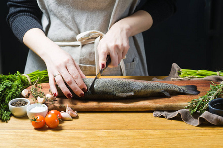 厨师在木制砧板上切生鳟鱼。 原料迷迭香柠檬西红柿大蒜盐胡椒。 黑色背景侧视图