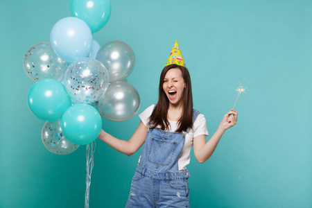 欢快的眨眼年轻女子戴着生日帽，手持燃烧的火花，用蓝色绿松石背景上分离的五颜六色的气球庆祝。 3.生日派对4.情感观念