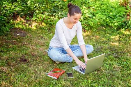 自由职业商业概念。 年轻女子坐在城市公园的绿草草坪上，用笔记本电脑工作。 生活方式真实，坦率的学生女孩在户外学习。 流动办公室