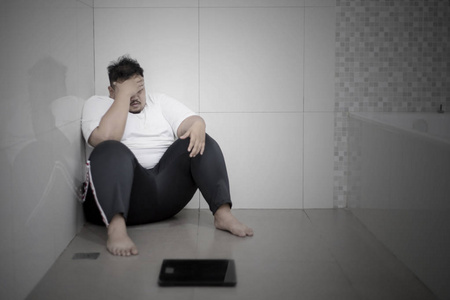 年轻的胖子在浴室里坐着秤,看上去很沮丧. 减肥失败概念照片