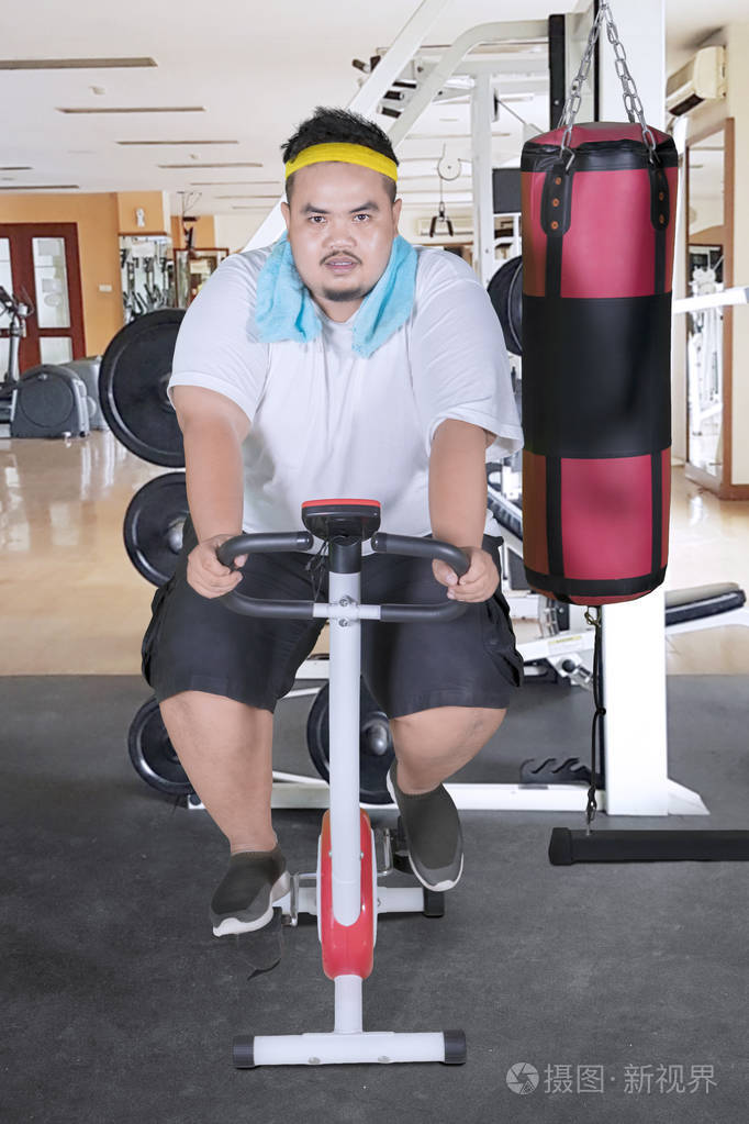 一位亚洲胖子穿着运动服,在健身房里骑运动自行车减肥的肖像