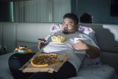 不健康的生活方式概念亚洲肥胖男子睡觉前在床上看电视时吃垃圾食品