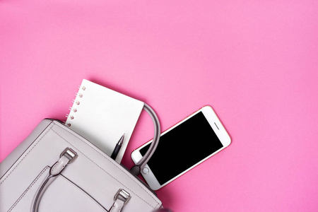 灰色的女性包和粉红色背景上的智能手机笔记本。 从上面看。 复制空间