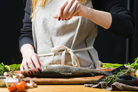 厨师在木制砧板上盐生鳟鱼。 原料迷迭香柠檬西红柿大蒜盐胡椒。 黑色背景侧视图