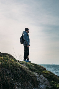 带着背包的男人旅行者站在草丛中的岩石悬崖上, 看着海景, 自由冒险的概念