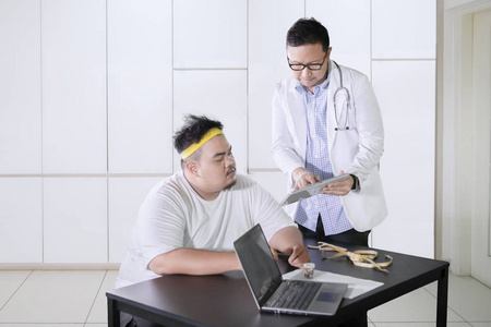年轻的男医生在诊所里和他肥胖的病人讨论，同时在药片上显示医疗结果