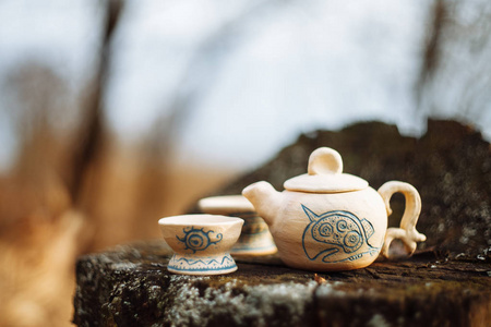泥白色茶壶，蓝色图案和两个饮酒碗在森林里的树桩。 手工。 秋季摄影