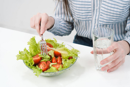 一个白色背景的年轻女孩坐在桌旁, 吃着一份绿色沙拉, 上面有西红柿和叉子, 喝着杯子里的水。健康食品