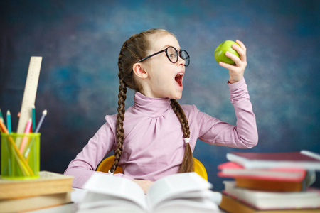 聪明的小学生长辫子在桌子上做学校活动. 孩子拿着绿苹果戴眼镜.