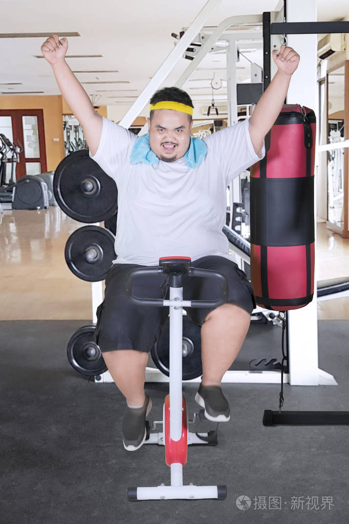 一个胖子在健身房里举起双手时用一辆表情愉快的运动自行车