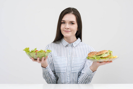 在白色背景的苗条的黑发女孩选择一盘快餐和汉堡包和健康食品沙拉