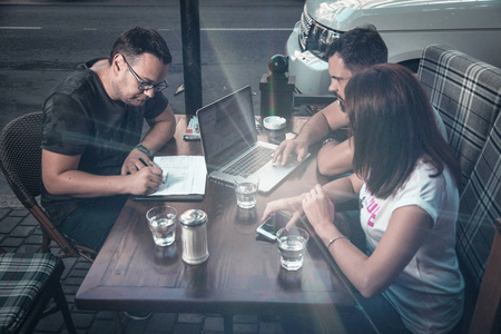 侧面视图和顶部。 这家由三位商人组成的公司正在餐桌上讨论咖啡，写在笔记本上，看着一台笔记本电脑，里面有水车钥匙，杂志，智能手机在