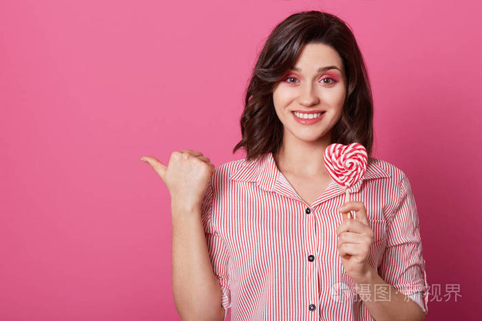 水平拍摄有吸引力的微笑妇女与高兴的表情点与拇指持有糖果的形式，心脏隔离在粉红色背景显示地方为您的广告内容。