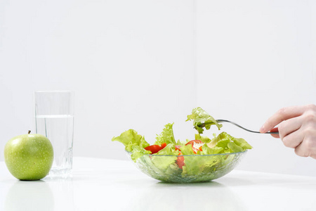 一个白色背景的年轻女孩坐在桌旁, 吃着一份绿色沙拉, 上面有西红柿和叉子, 喝着杯子里的水。健康食品