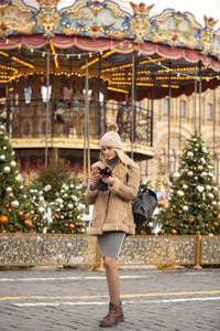 一位身穿羊皮大衣戴着针织帽子的戴着手机在圣诞庭院中央拍照的金发美女