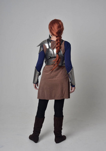 一个穿着中世纪战士服装和钢甲站立姿势的红头发女孩的全长肖像，在灰色工作室背景下回到相机上。