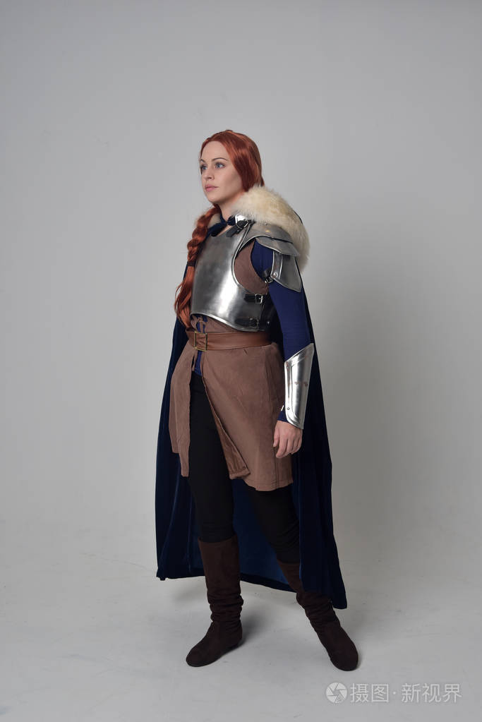 一个穿着中世纪战士服装蓝色天鹅绒斗篷和钢甲站立姿势的红头发女孩在