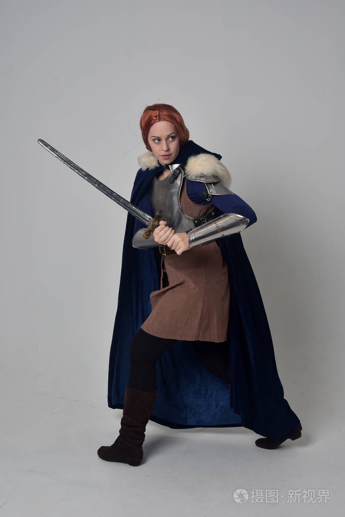 一个穿着中世纪战士服装,蓝色天鹅绒斗篷和钢甲站立姿势的红头发女孩