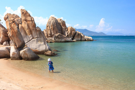 小女孩站在岩石海岸的水中，有热带植被，在海湾有沙滩