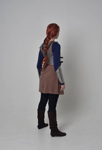 一个穿着中世纪战士服装和钢甲站立姿势的红头发女孩的全长肖像，在灰色工作室背景下回到相机上。