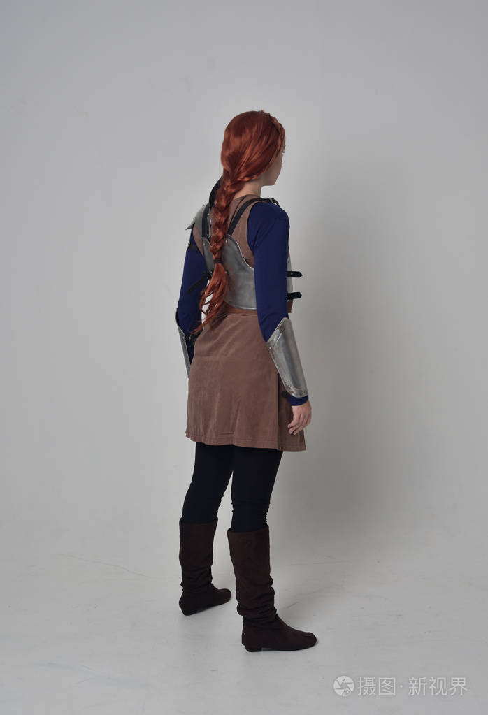 一个穿着中世纪战士服装和钢甲站立姿势的红头发女孩的全长肖像,在灰