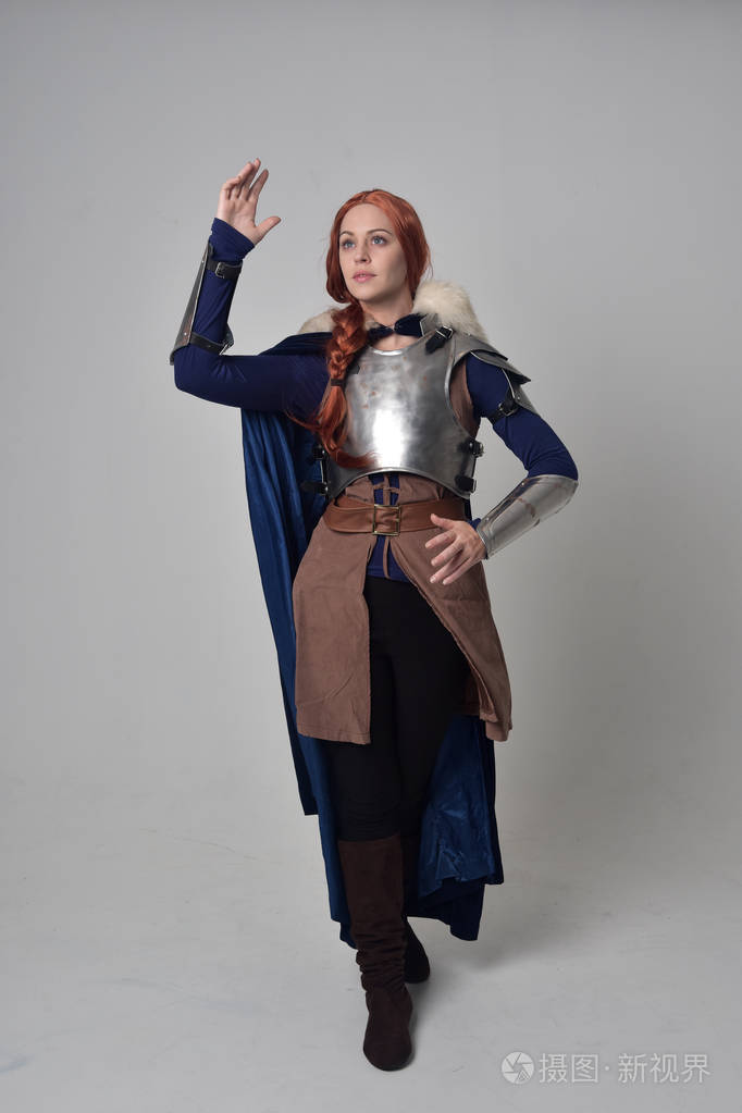 一个穿着中世纪战士服装,蓝色天鹅绒斗篷和钢甲站立姿势的红头发女孩
