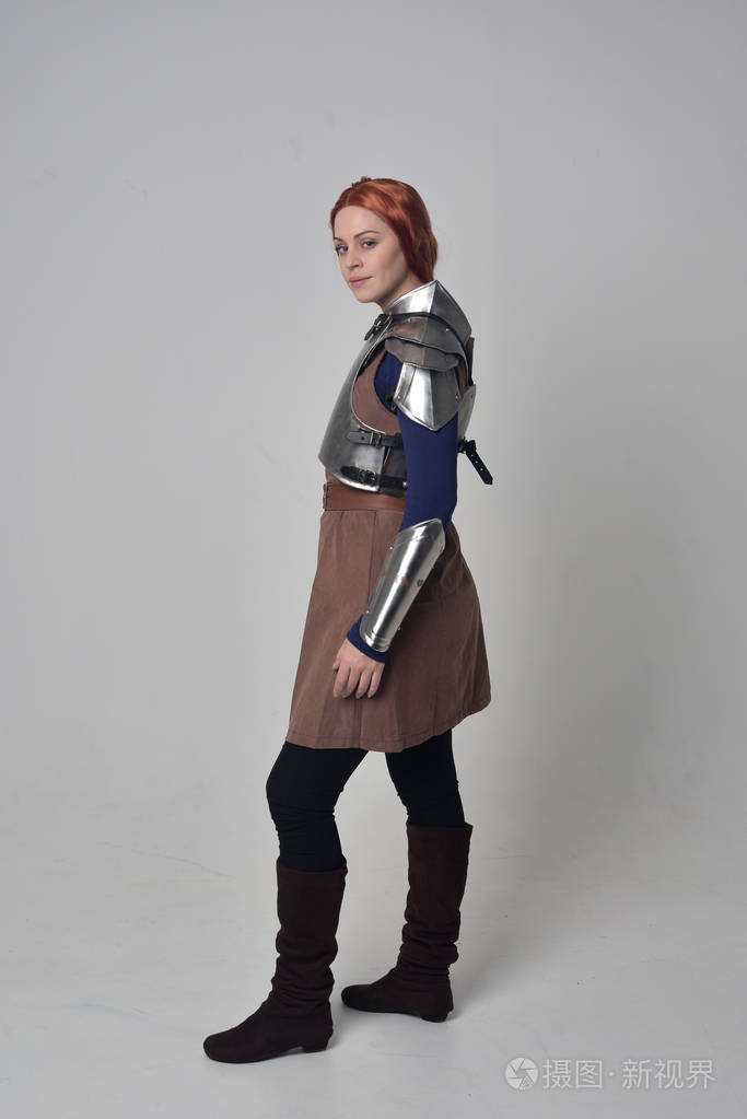 一个穿着中世纪战士服装和钢甲站立姿势的红头发女孩在灰色工作室背景