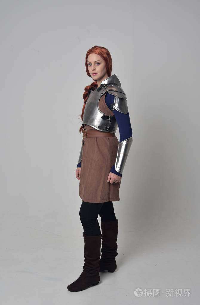 一个穿着中世纪战士服装和钢甲的红头发女孩的全长肖像站在灰色工作室