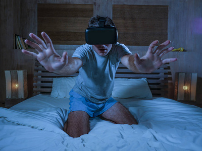年轻的兴奋和惊讶的男人，穿着VR虚拟现实护目镜设备，玩互联网3D模拟器游戏，享受令人兴奋和令人震惊的视觉体验，家中的床。