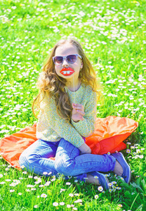 女孩坐在草地上的草地, 绿色的背景。孩子摆着眼镜纸板微笑的嘴唇在草地上拍照。幽默的概念。女孩在愉快的面孔花费休闲户外