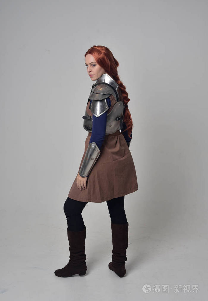 一个穿着中世纪战士服装和钢甲的红头发女孩的全长肖像,站在灰色工作