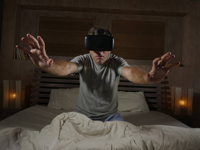 年轻的兴奋和惊讶的男人，穿着虚拟现实护目镜设备，玩互联网3D模拟器游戏，享受令人兴奋和震惊的视觉体验，家中的床。
