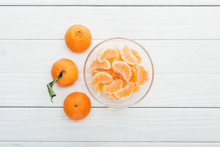 玻璃碗中的去皮橘子切片和木制白色背景上的全熟橘子的顶部视图。