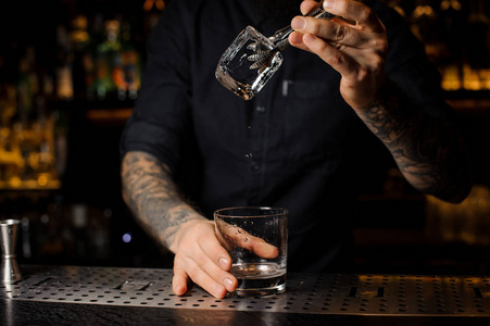 酒保在杯子里的饮料里加入一个大冰块，柜台上有镊子
