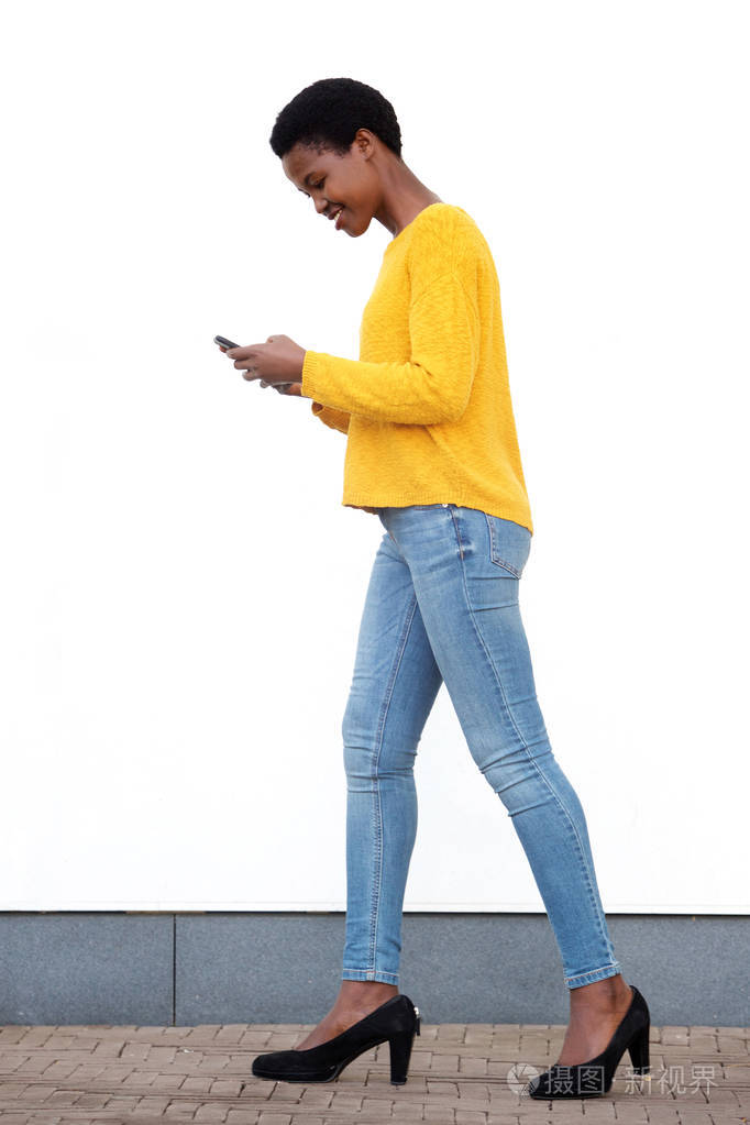 年轻的黑人妇女在白色的墙边用手机走路的全身侧面肖像
