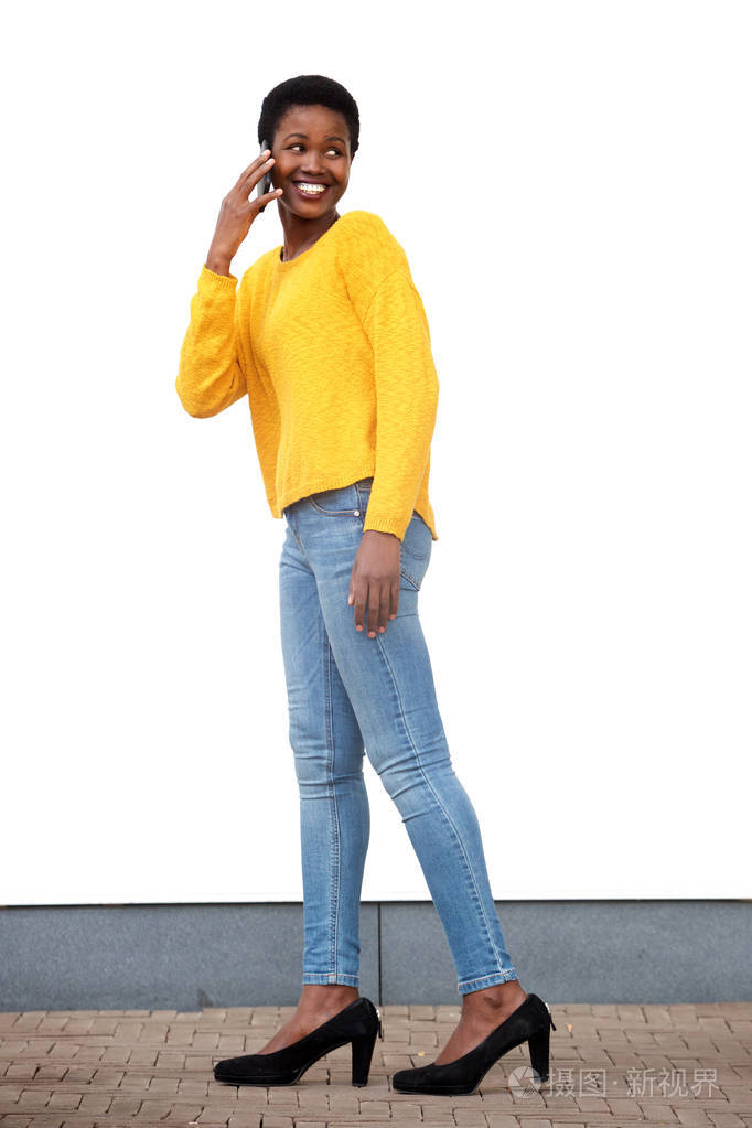 美国黑人年轻女子在白色墙边用手机走路的全身侧面肖像
