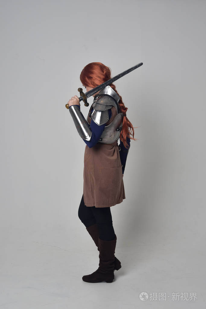 一个穿着中世纪战士服装和钢甲的红头发女孩的全长肖像,手持一把剑.