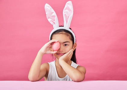 摄影棚拍摄了一个快乐的小女孩，她戴着兔子的耳朵，在粉红色的背景上举起一个五颜六色的复活节彩蛋