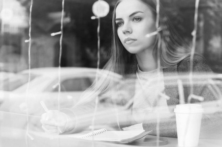 单色黑白肖像一个有吸引力的年轻记者女孩与笔和记事本在展示后面的咖啡馆。通过窗户玻璃反射的非对比度视图