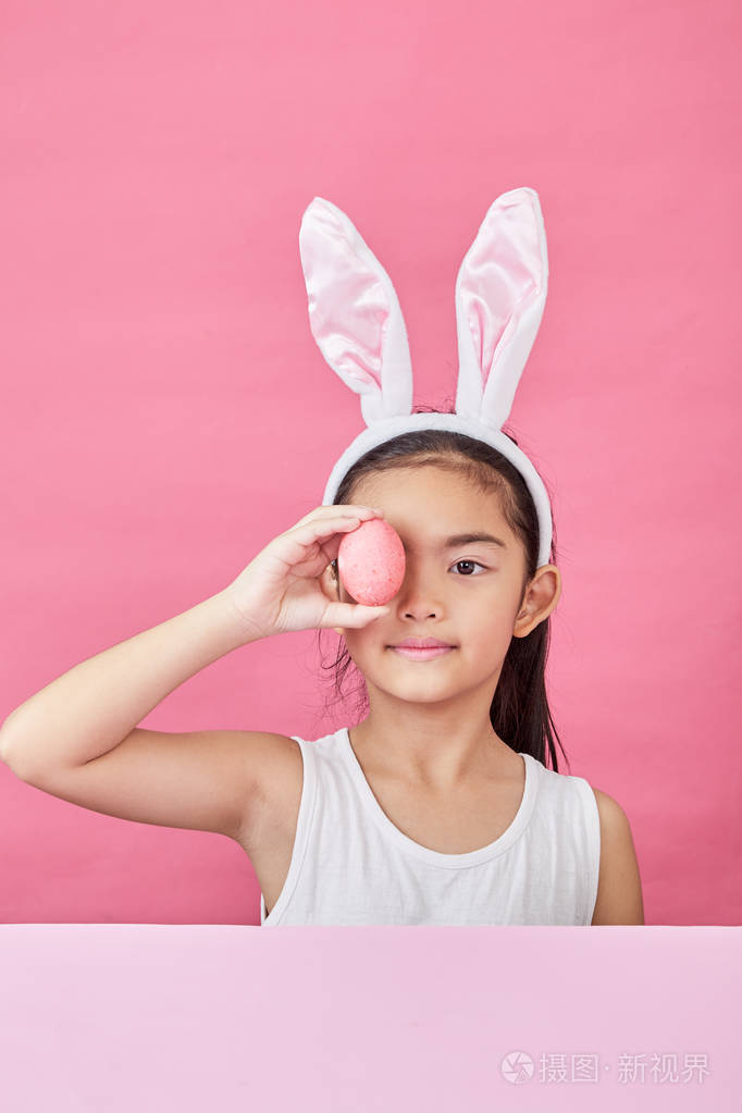摄影棚拍摄一个快乐的小女孩戴着兔子耳朵,在粉红色的背景上举起一个