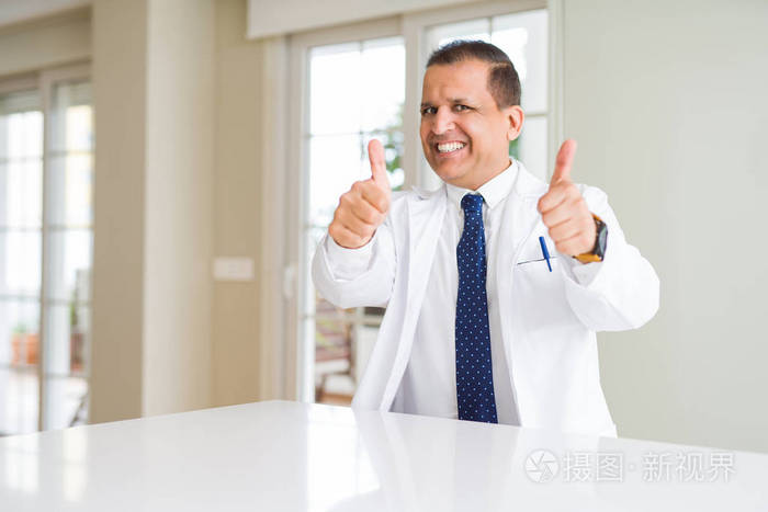中年医生男子穿着医疗外套在诊所批准做积极的手势，用手竖起大拇指，微笑和快乐的成功。 看着相机赢家的手势。