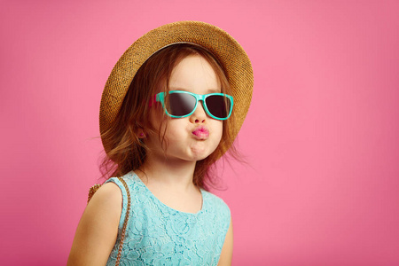 美丽的小女孩穿着时髦的衣服, 戴着草帽, 太阳镜和蓝色的衣服, 显示鸭脸, 站在粉红色孤立