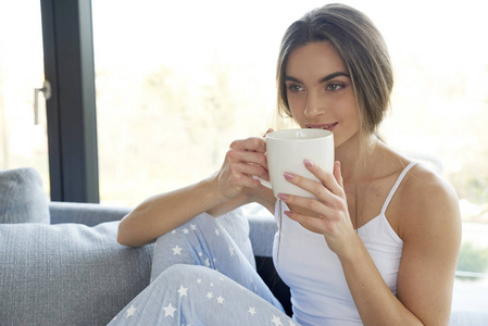 特写照片拍摄的年轻女子穿着睡衣，喝着早茶，在家里的沙发上放松。