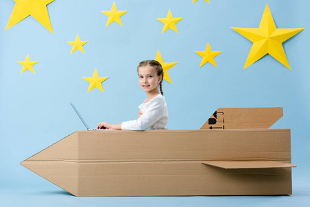 快乐的孩子，带着大纸板火箭，用笔记本电脑，蓝色背景