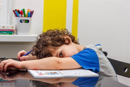 疲惫的小男孩坐在桌旁, 胳膊上着头, 一边做作业, 一边在笔记本上写字。学校大量作业的概念