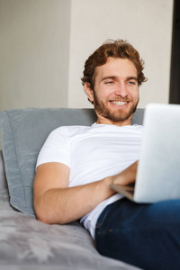 一个英俊的年轻胡子男子在家里沙发上使用笔记本电脑的图像。