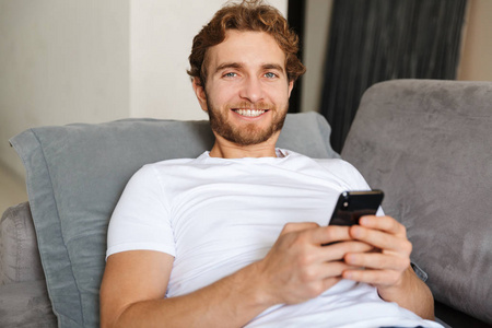 一个英俊的年轻留胡子的男人在家里用手机在沙发上的形象。