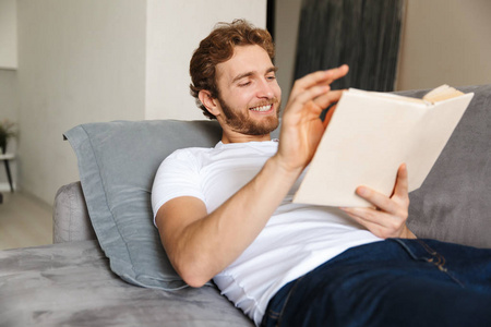 一个英俊的年轻胡须男在沙发上在家看书的形象。