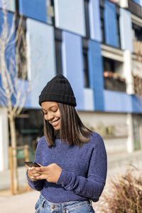 在城市街道上戴帽子的幸福女人的肖像，同时在户外使用技术，手持手机。 她二十出头就是黑人