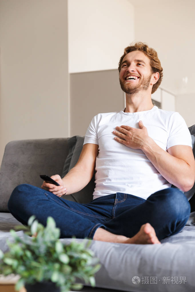 一个英俊快乐的年轻人在家沙发上拿着遥控器看电视的照片